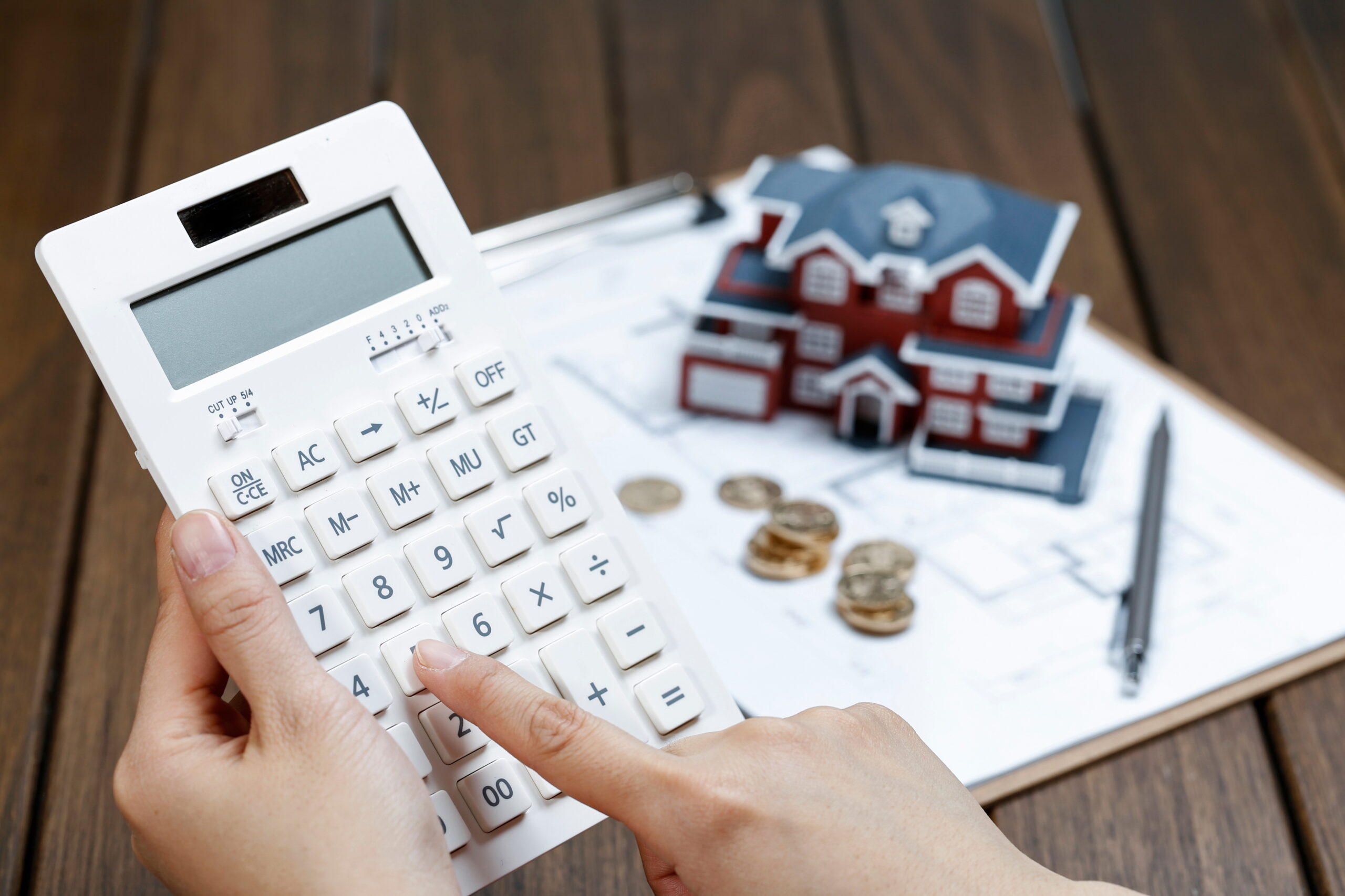 Close-up of hands using a calculator beside architectural plans, coins, and a house model—representing financial analysis for apartment building decisions.