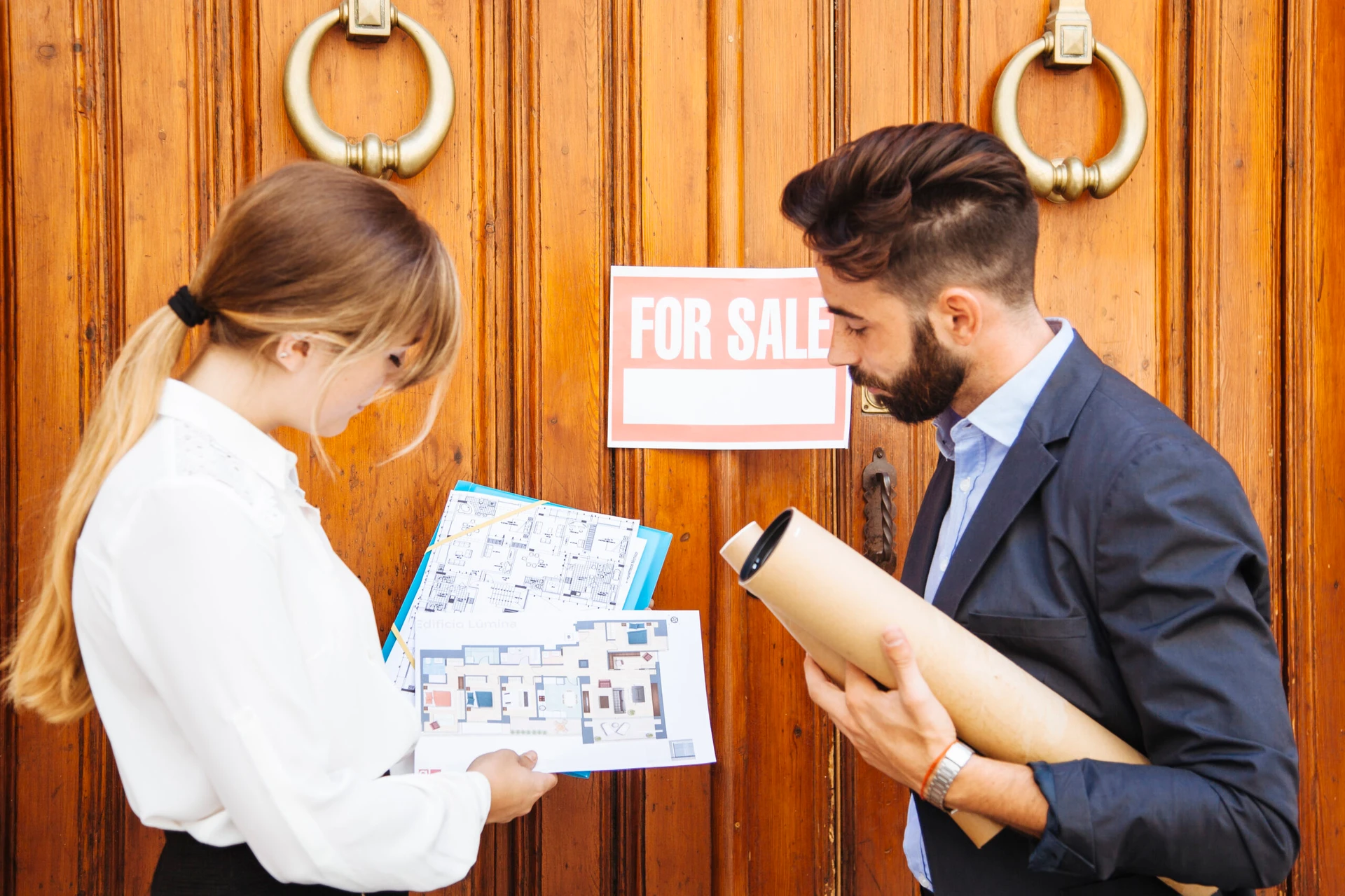 Two professionals reviewing architectural plans in front of a large apartment building door with a "For Sale" sign, symbolizing a decision between refinancing or selling.