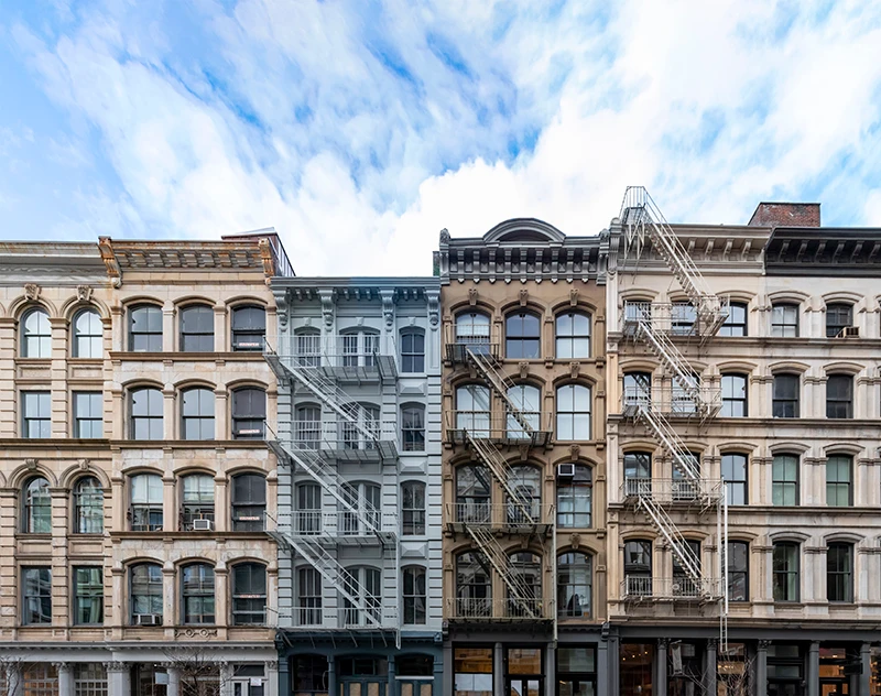Row of ornate 19th-century buildings with arched windows, decorative facades, and fire escapes under a partly cloudy sky.