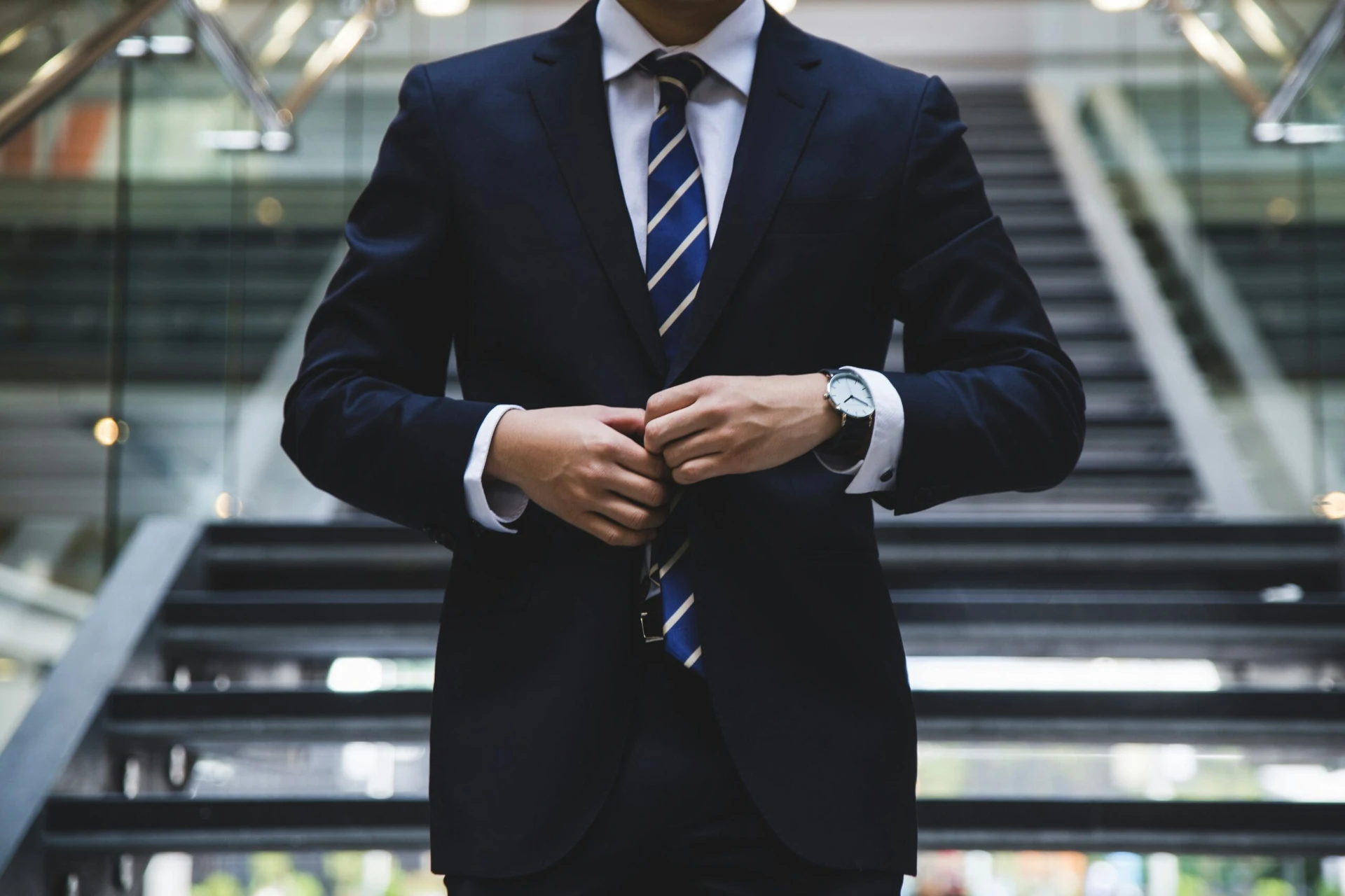 Business professional in a navy suit adjusting his jacket, standing on a modern indoor staircase.